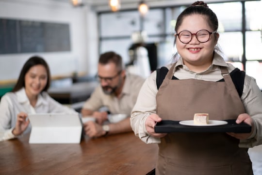 Young waitress with Down syndrome smiles at the camera at work while serving a piece cake on a tray.